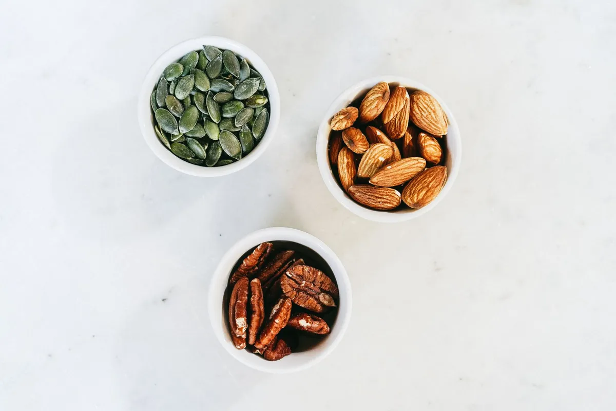 Small bowls of green pumpkin seeds, almonds and pecans seen from above on a white table